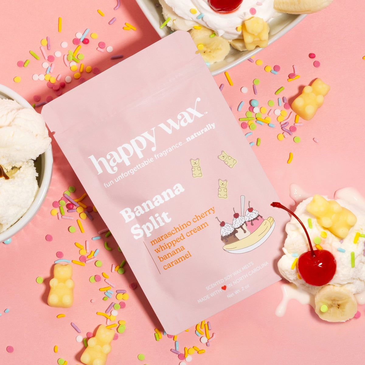 Pink package of Banana Split wax melts surrounded by yellow wax cubes, sprinkles, and banana slices on pink background.