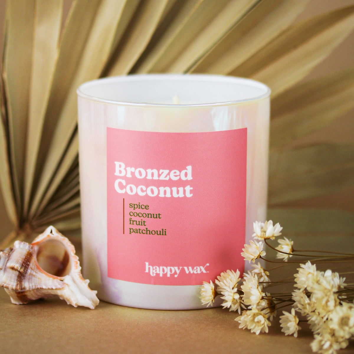 White cylindrical candle with pink label reading "Bronzed Coconut" and scent notes, single wick visible, surrounded by shells