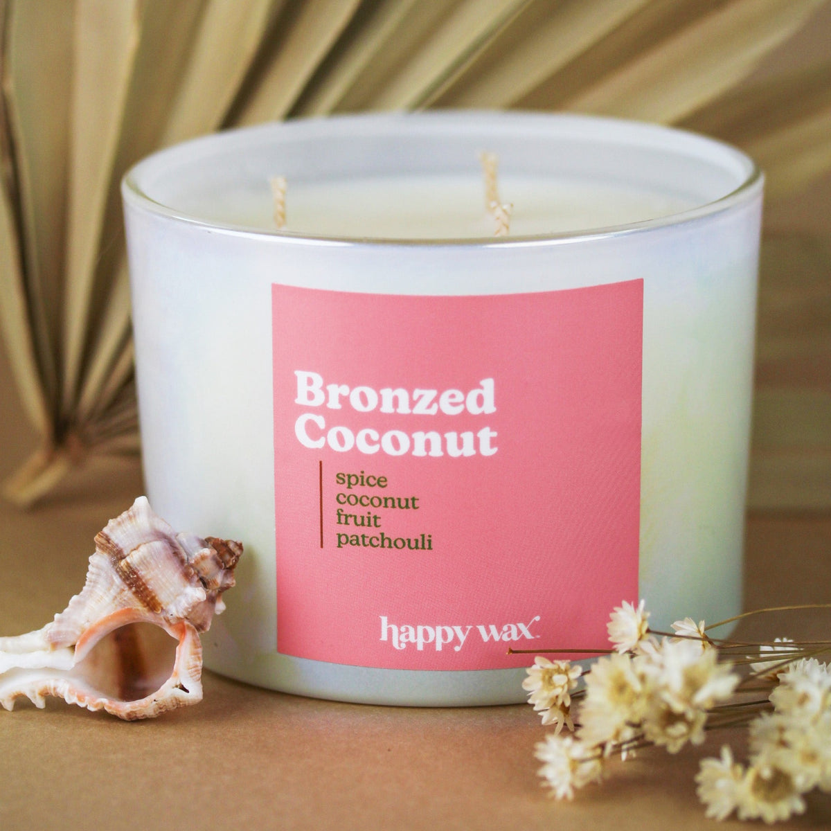 White three-wick candle with pink label reading "Bronzed Coconut" by happy wax, surrounded by seashell and dried flowers.
