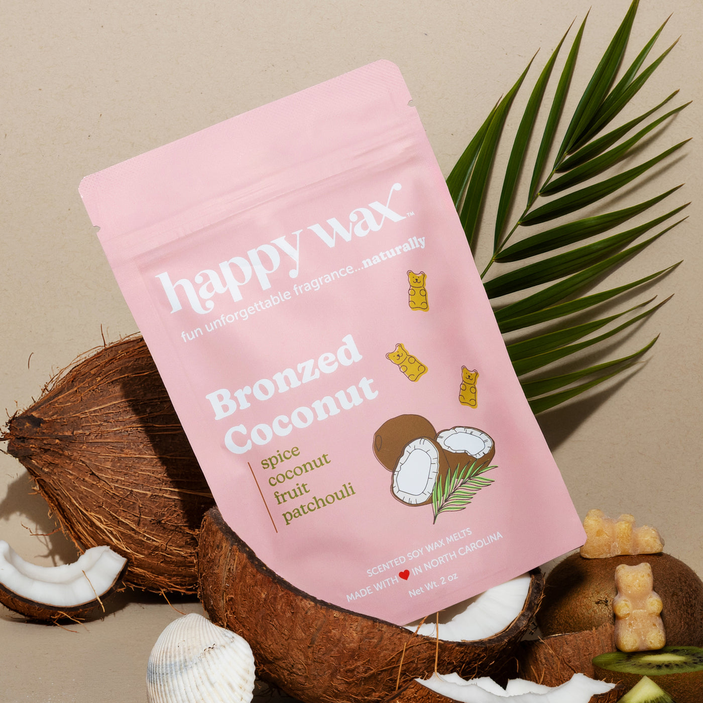 Pink pouch of Bronzed Coconut wax melts surrounded by coconut halves, palm fronds, and gummy bears on beige background.