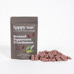 Brown pouch of Happy Wax wax melts labeled "Bronzed Peppercorn & Cardamom" with scattered rose-colored wax melts displayed in