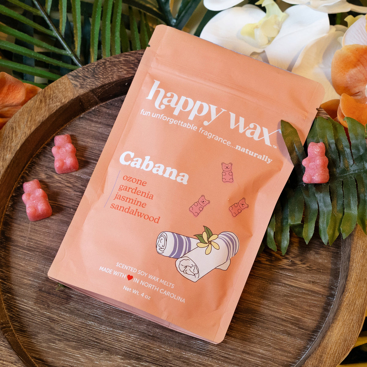 Peach-colored Happy Wax pouch with Cabana scent surrounded by pink wax melts on wooden tray with greenery.