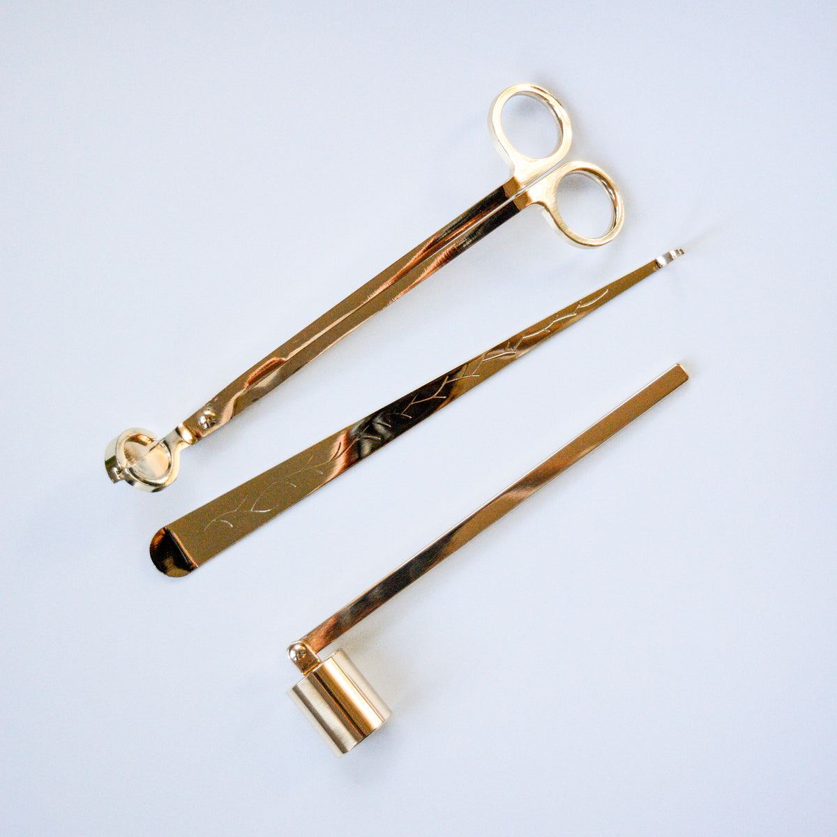 Three gold-toned candle care tools: scissors, wick trimmer, and candle snuffer arranged on white background.
