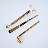 Three gold-toned candle care tools: scissors, wick trimmer, and candle snuffer arranged on white background.
