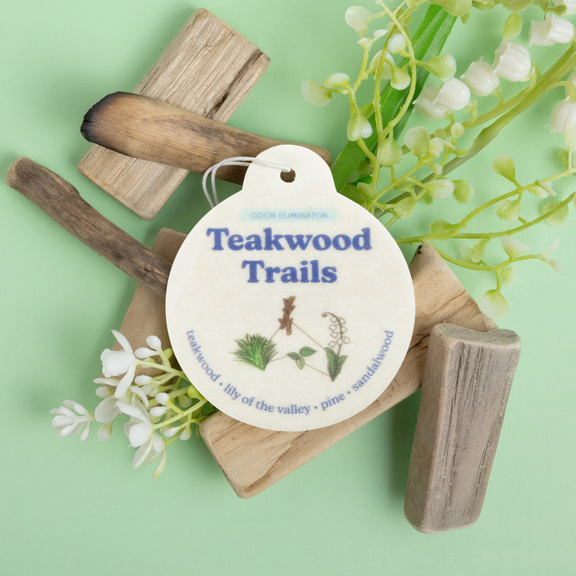 White oval car charm labeled "Teakwood Trails" with wooden pieces and white flowers on green background.