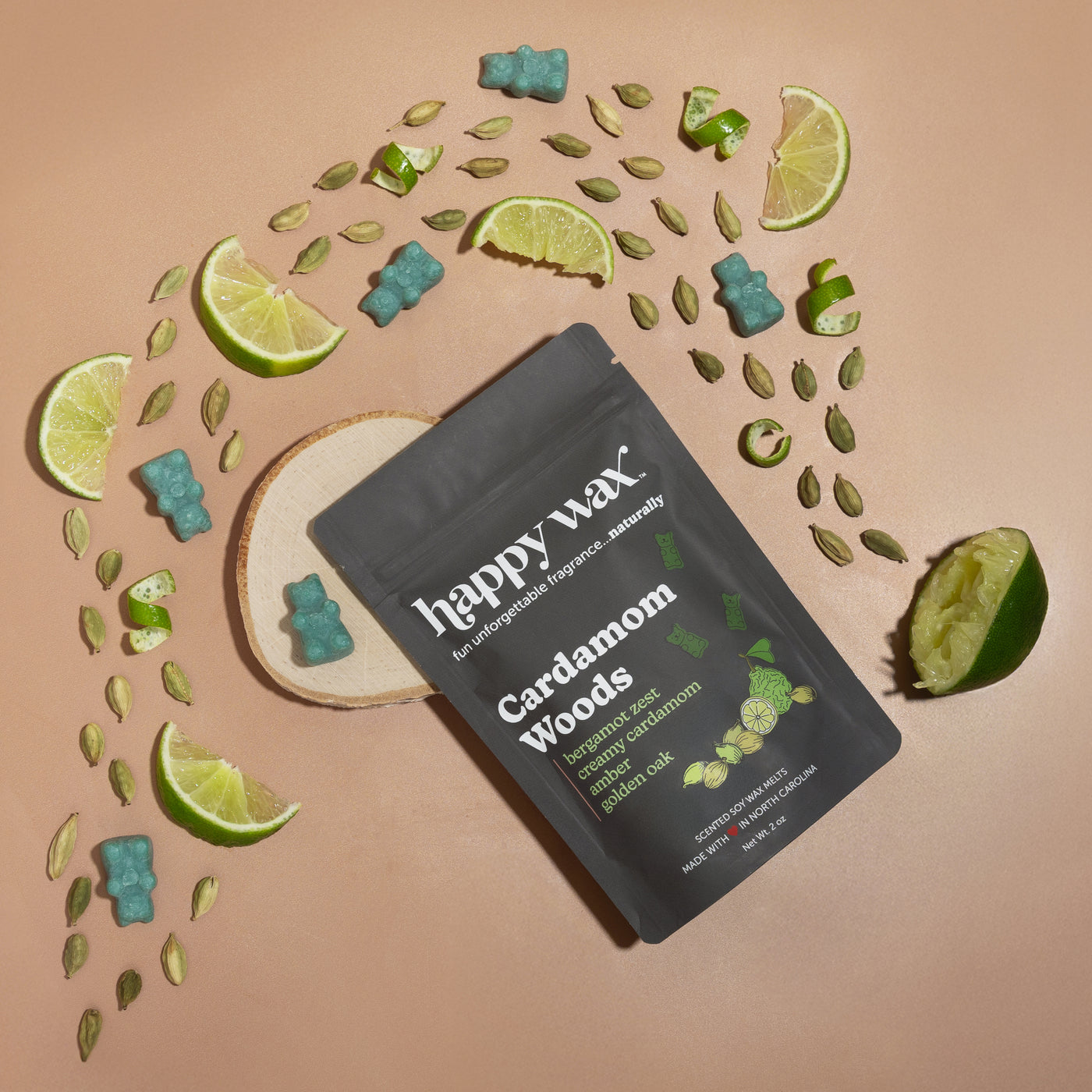 Black pouch of Cardamom Woods wax melts surrounded by cardamom pods, lime slices, and teal wax melts on beige background.