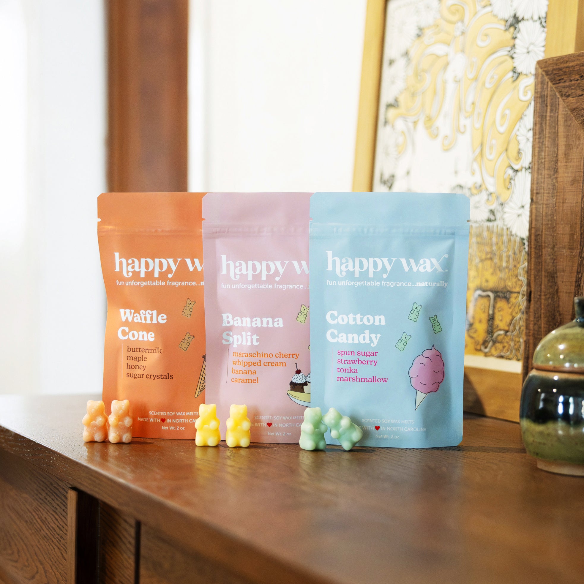 Three Happy Wax scent packets in orange, pink, and blue with wax melts displayed in front on a wooden table.
