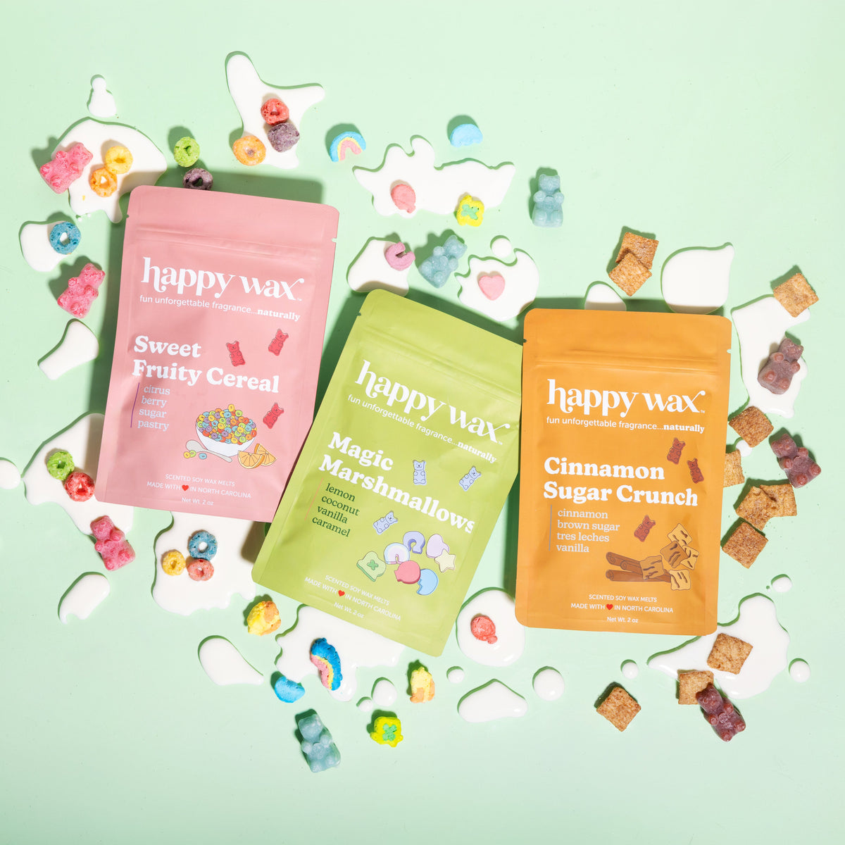 Three Happy Wax cereal-scented candle pouches in pink, green, and orange with colorful wax melts scattered around on pale gre