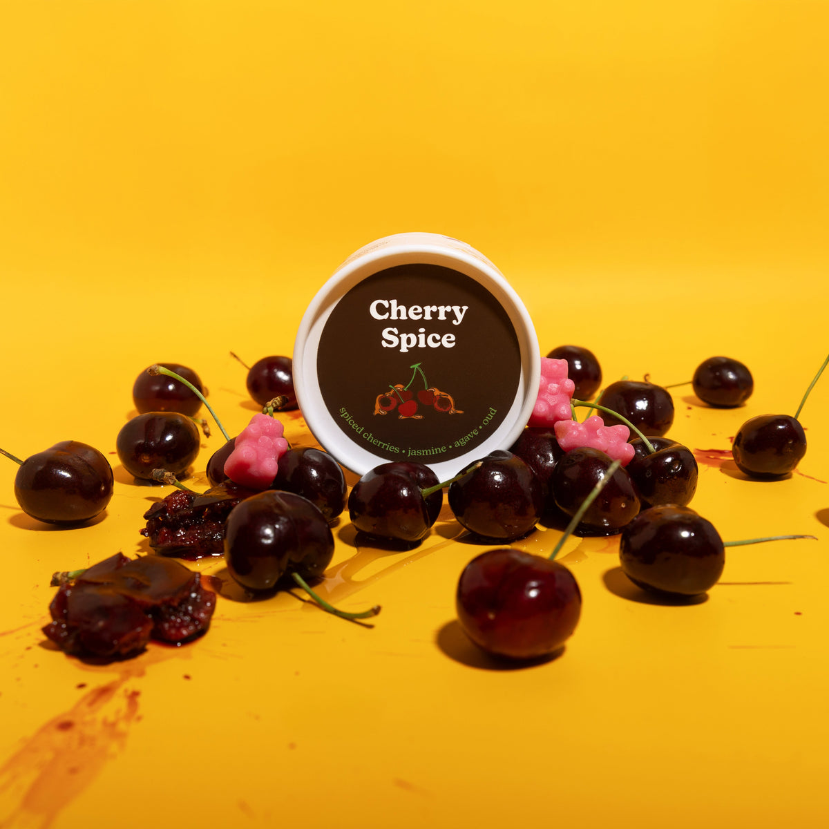 Round wax melt container with brown label surrounded by dark cherries and pink peppercorns on yellow background.