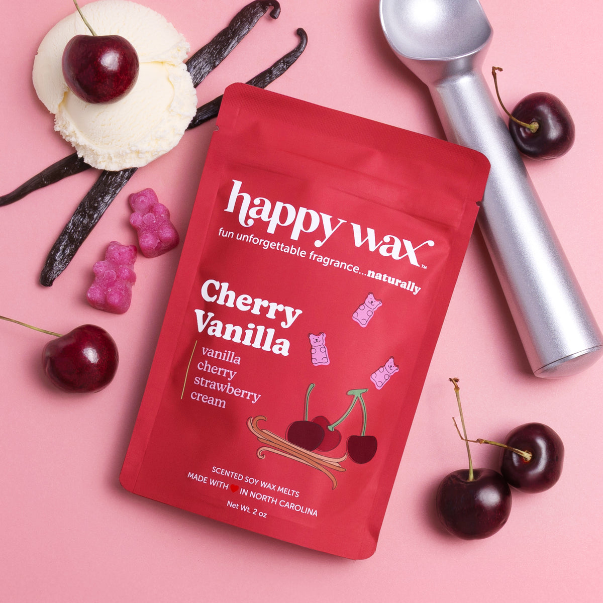 Red happy wax packet with cherry vanilla scent surrounded by cherries, vanilla beans, and a silver melter on pink background.