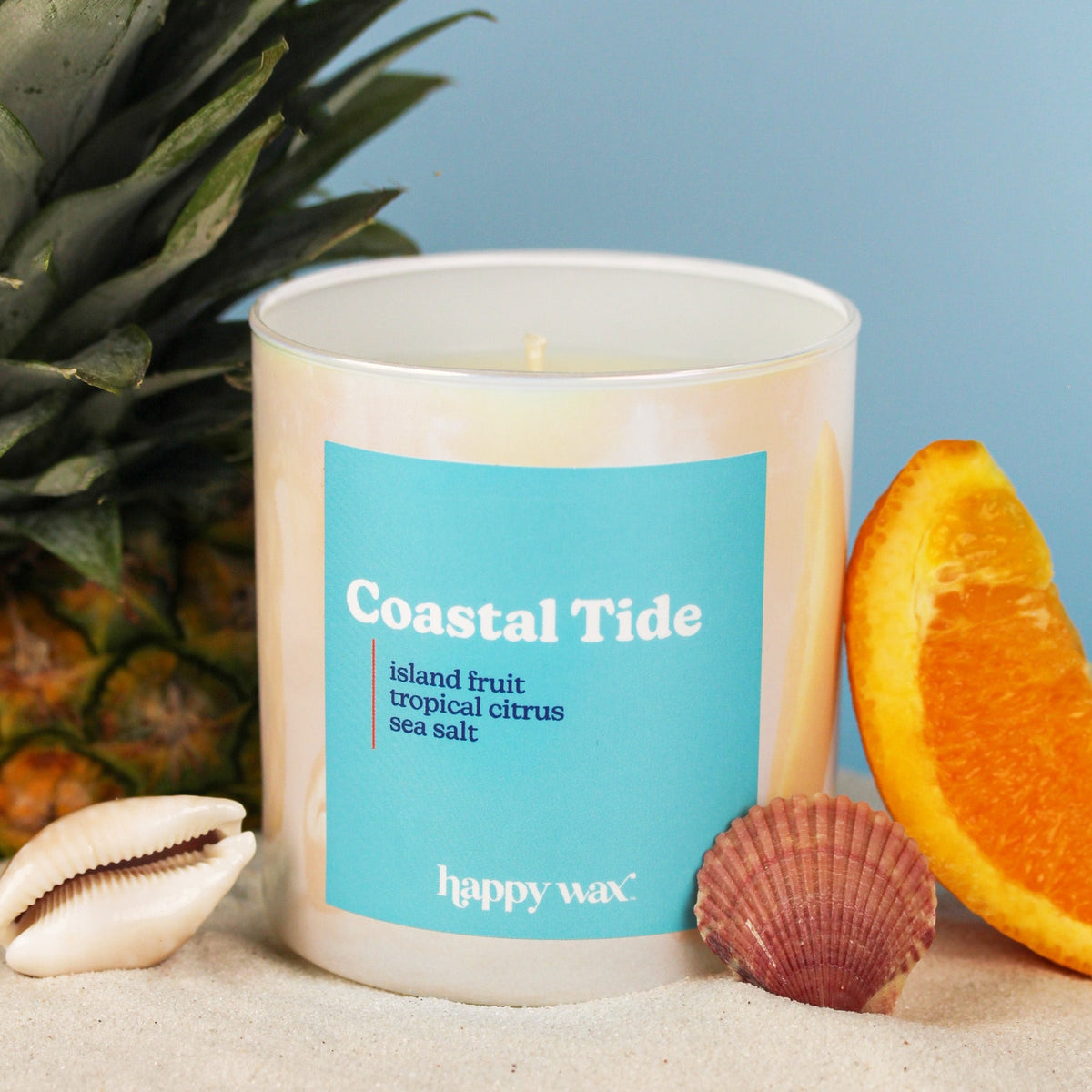 Cream glass candle with turquoise label, single lit wick, surrounded by pineapple leaves, orange slice, seashell, and sand.