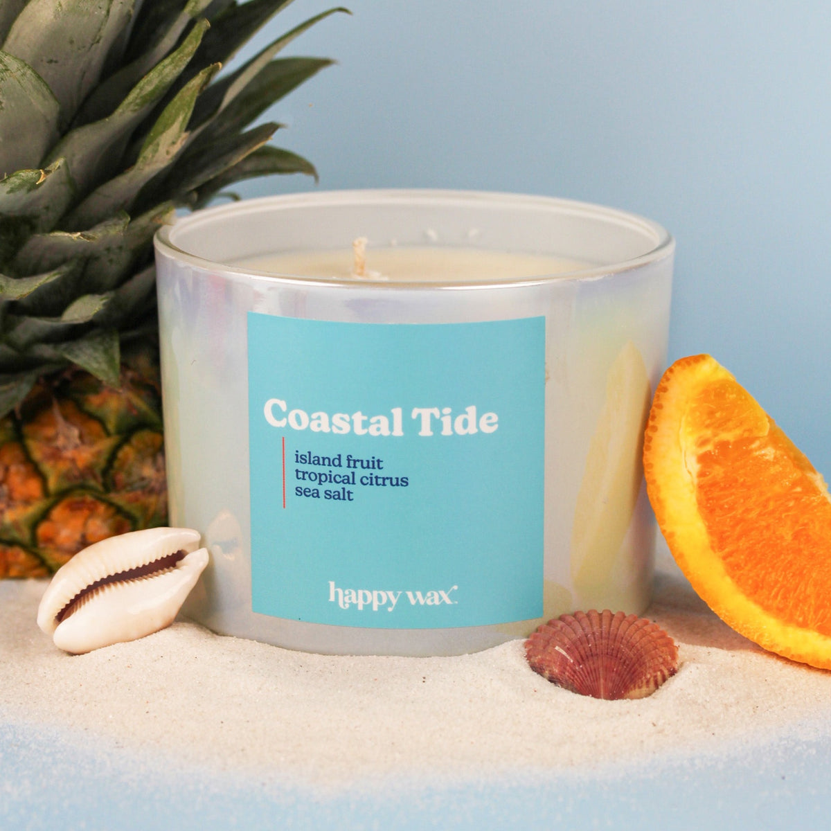 Three-wick candle with turquoise label surrounded by pineapple, orange slice, seashells, and sand.