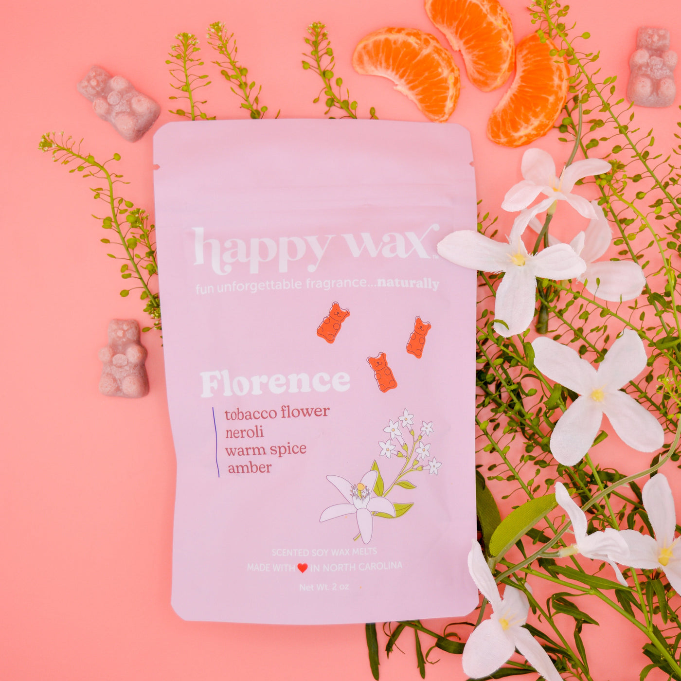 Pink pouch of Florence wax melts surrounded by white flowers, orange citrus slices, green foliage, and scattered wax melts on