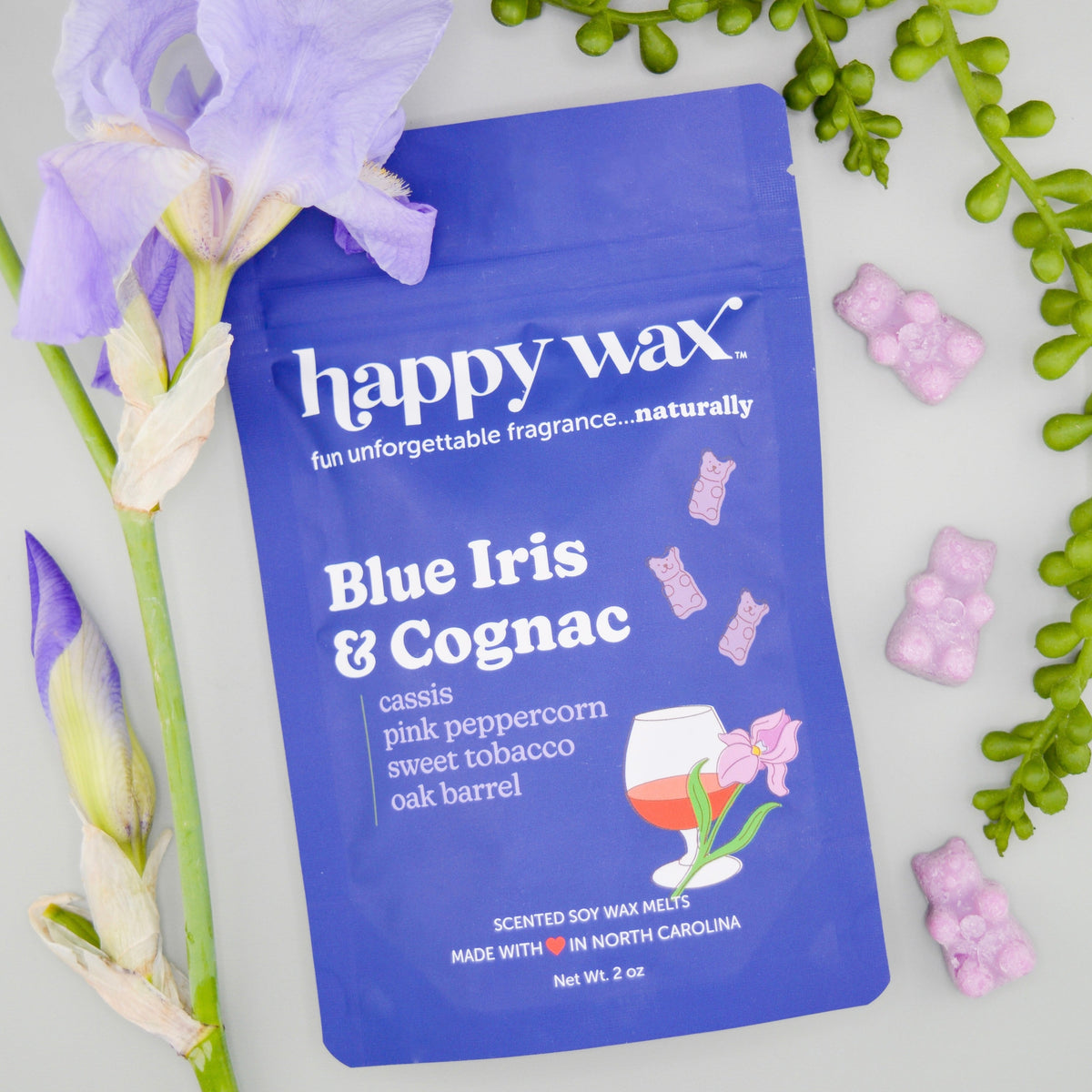 Blue pouch of Happy Wax brand wax melts with pink cube melts displayed alongside purple iris flower and green foliage.