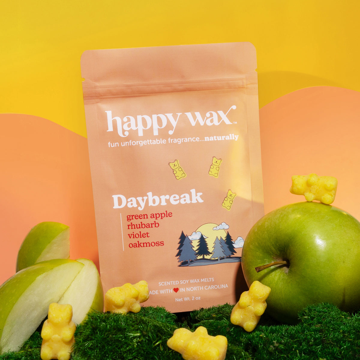Tan kraft pouch of Happy Wax Daybreak soy wax melts surrounded by green apples, gummy bears, and moss against yellow and oran