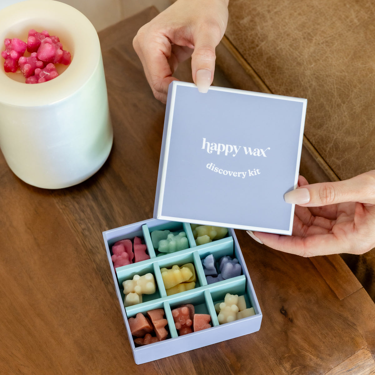 Hand holding open Happy Wax discovery kit box displaying nine compartments of colored wax melts next to white warmer bowl wit