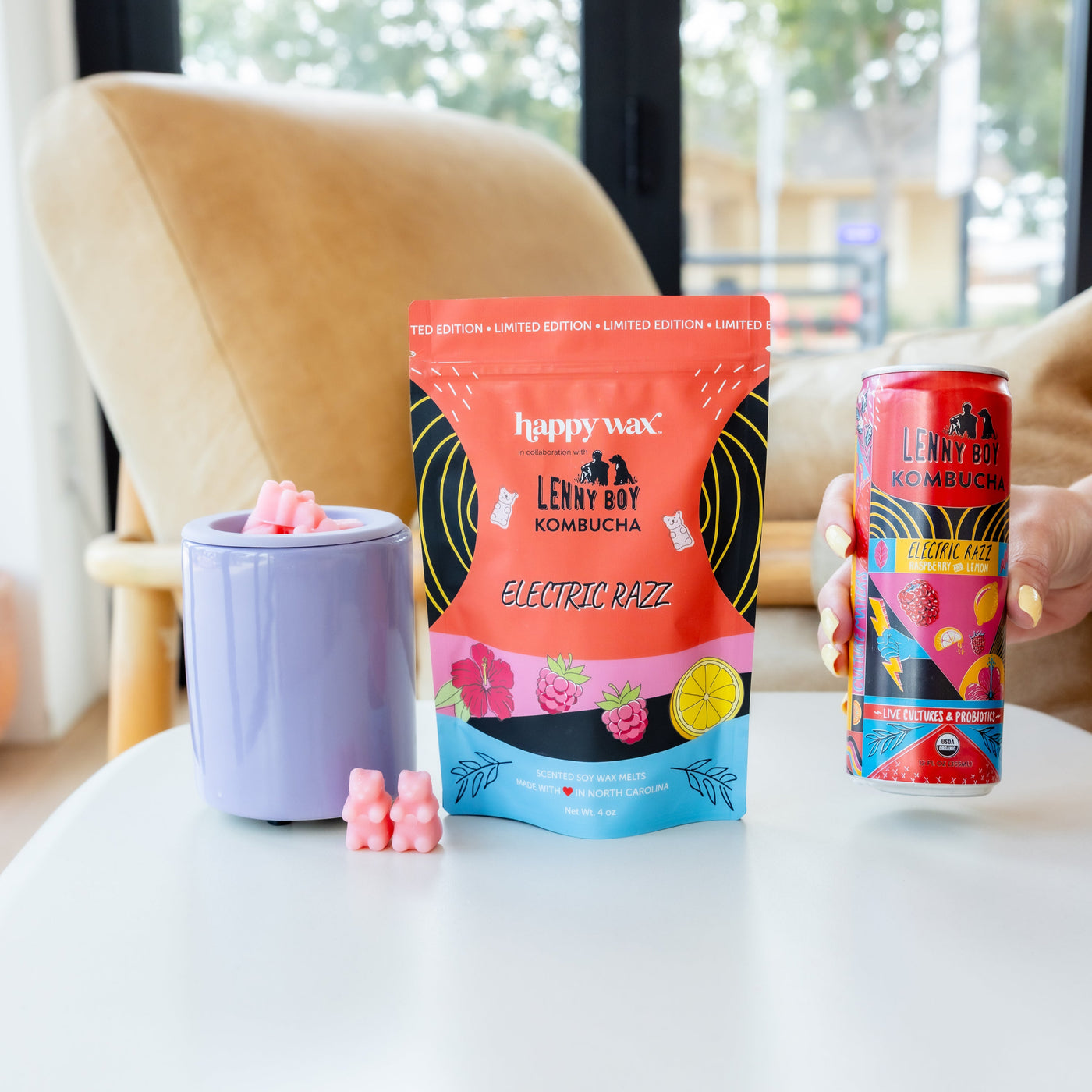Pink wax melts in lavender warmer with red Happy Wax packaging and matching kombucha beverage can displayed on white surface.