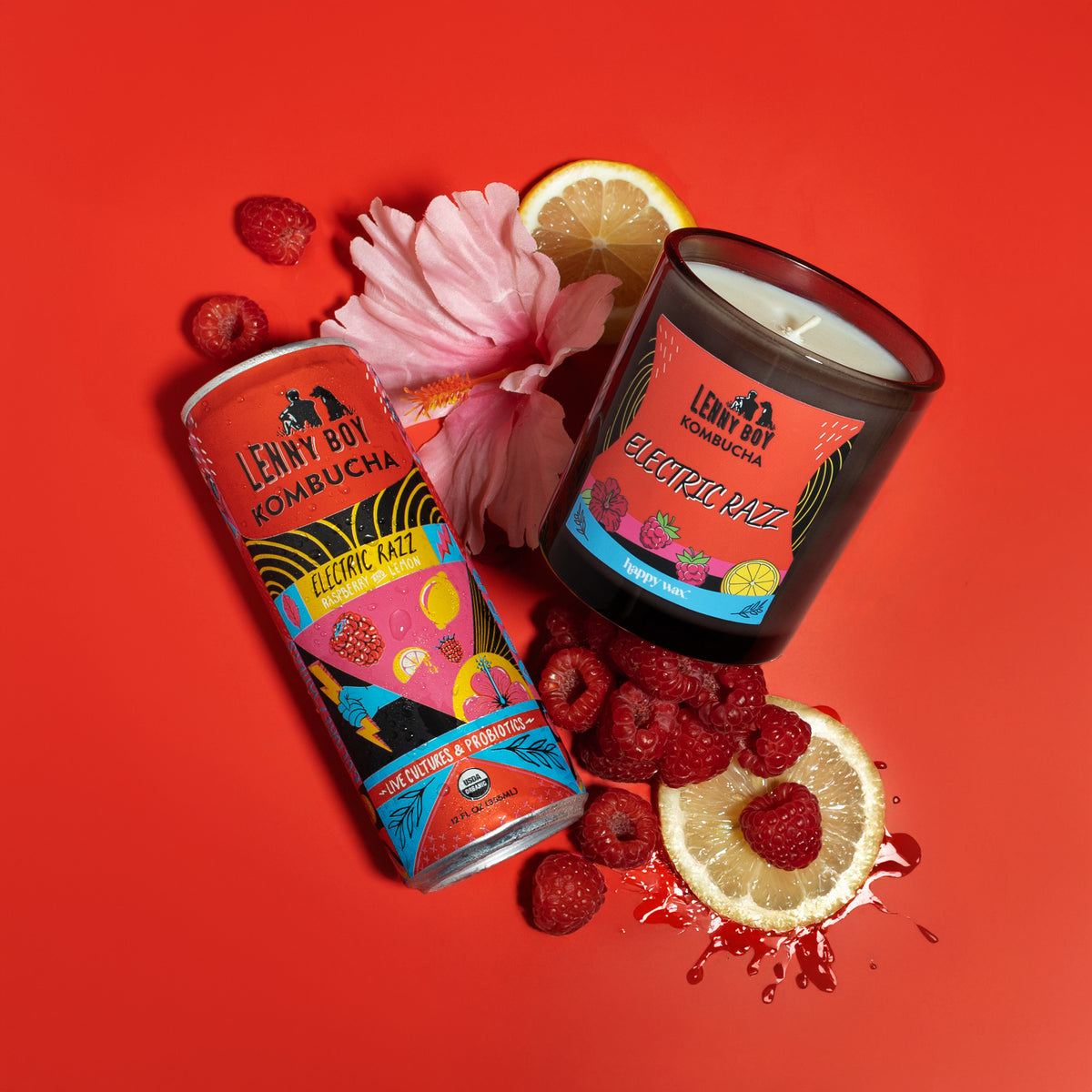 Dark glass candle jar with white wick surrounded by raspberries, lemon slices, and pink flower on red background.