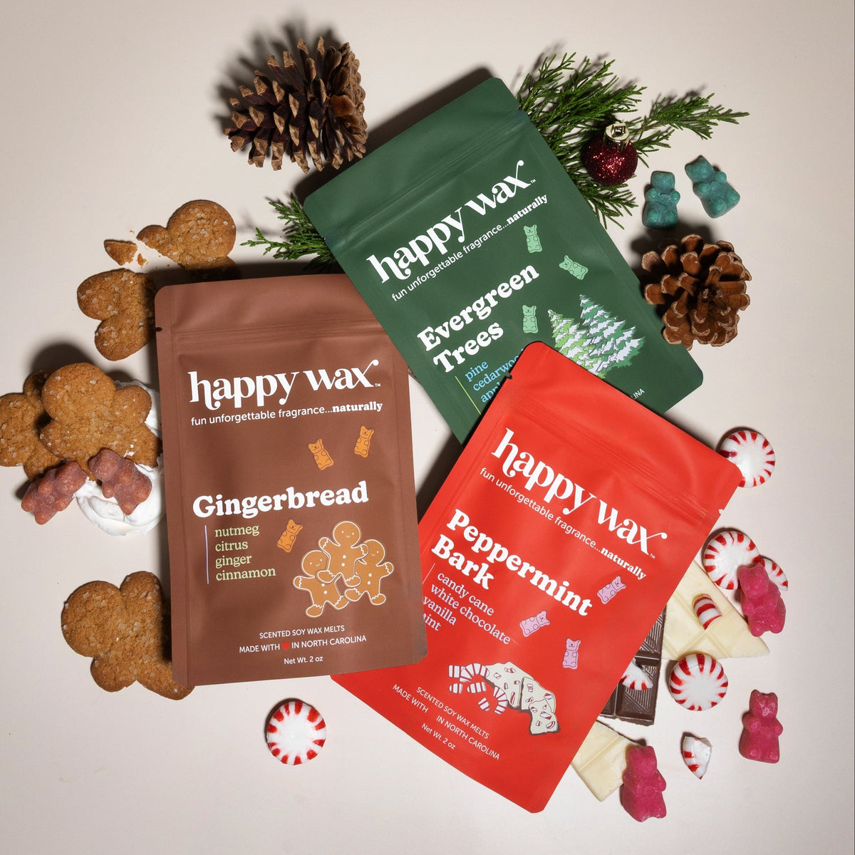 Three Happy Wax scent packets in brown, green, and red with gingerbread, evergreen, and peppermint flavors surrounded by pine
