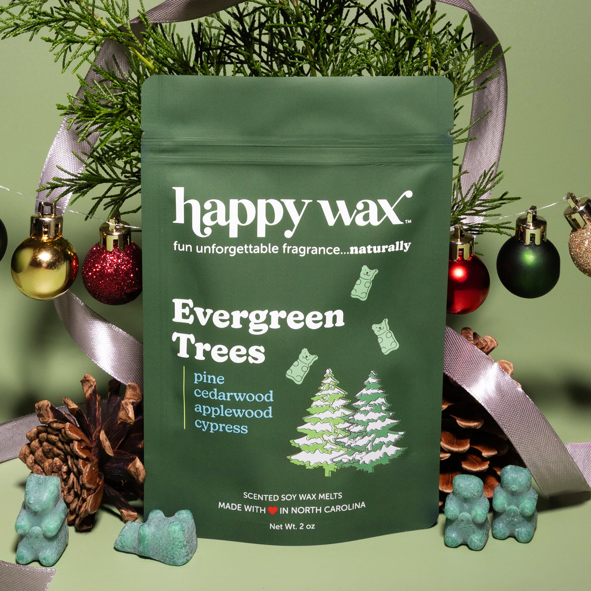 Green pouch of Happy Wax evergreen-scented soy wax melts surrounded by holiday ornaments, pinecones, and decorative items.
