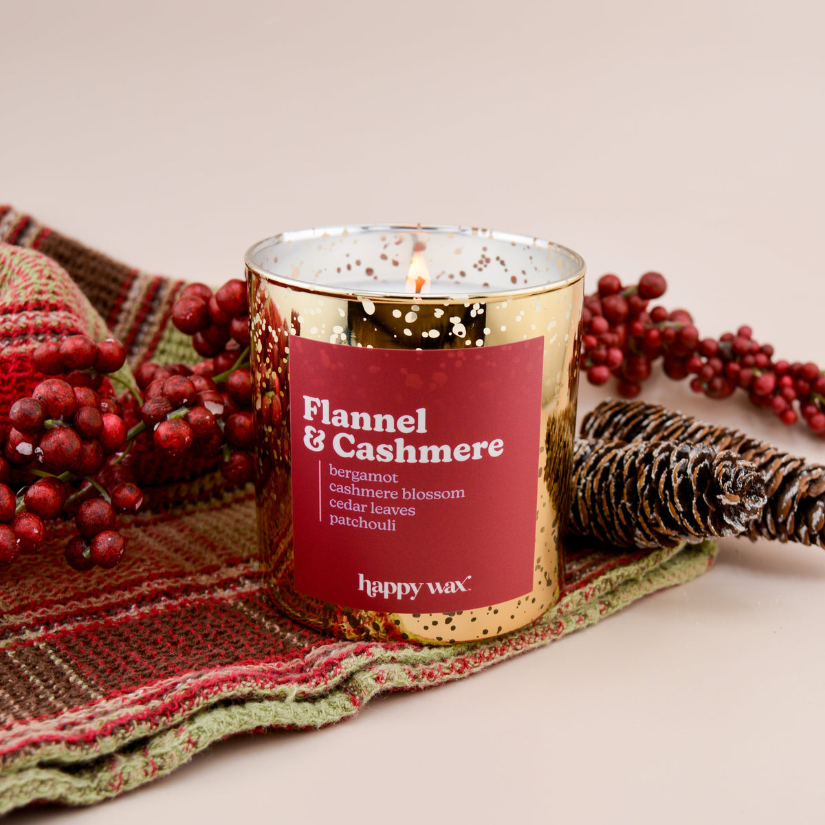Glass candle with red label reading "Flannel & Cashmere" surrounded by plaid fabric, red berries, and woven decorative elemen