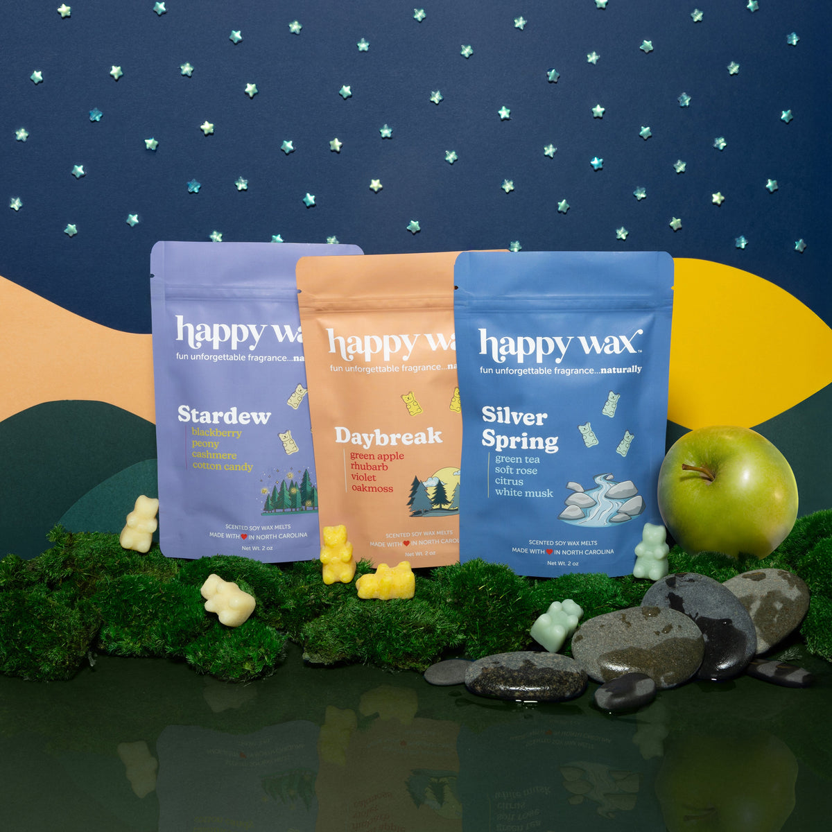 Three Happy Wax scented wax melt pouches in purple, tan, and blue with wax melts and natural elements displayed against a sta