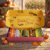 Yellow subscription box labeled "Golden Acre" with farm illustrations, containing packaged food items and a green mug.