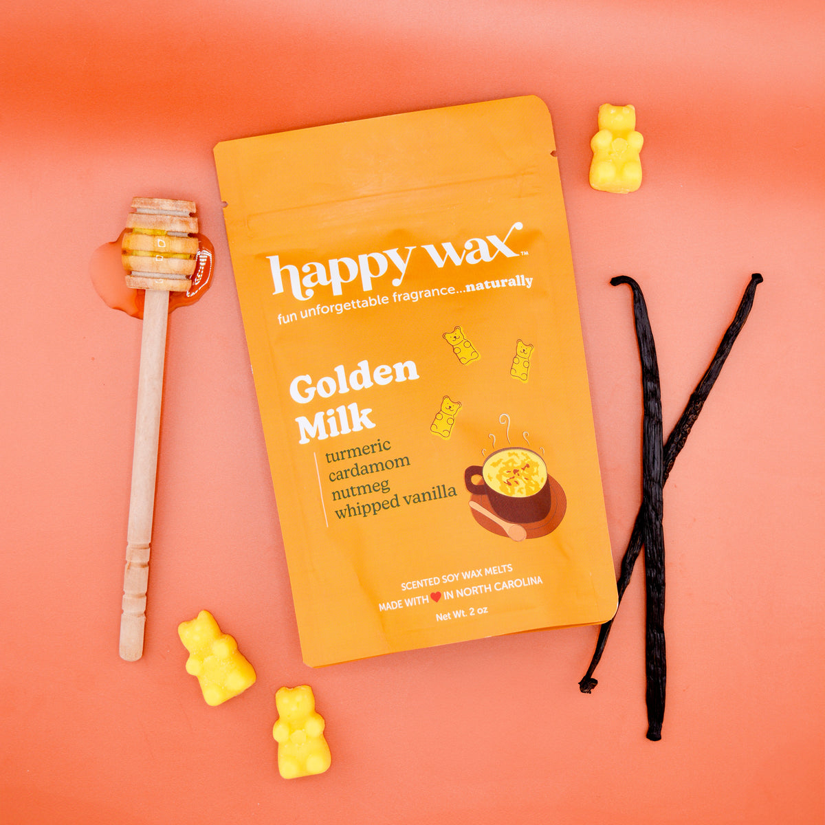 Golden Milk wax melt pouch surrounded by yellow bear-shaped melts, vanilla pods, and wooden stirring stick on coral backgroun