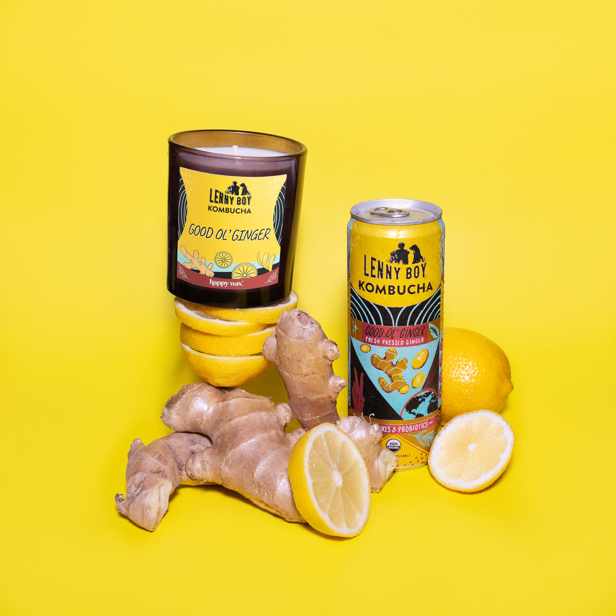 Amber glass candle with yellow label reading "Good Ol' Ginger," surrounded by fresh ginger root, lemons, and a kombucha can o