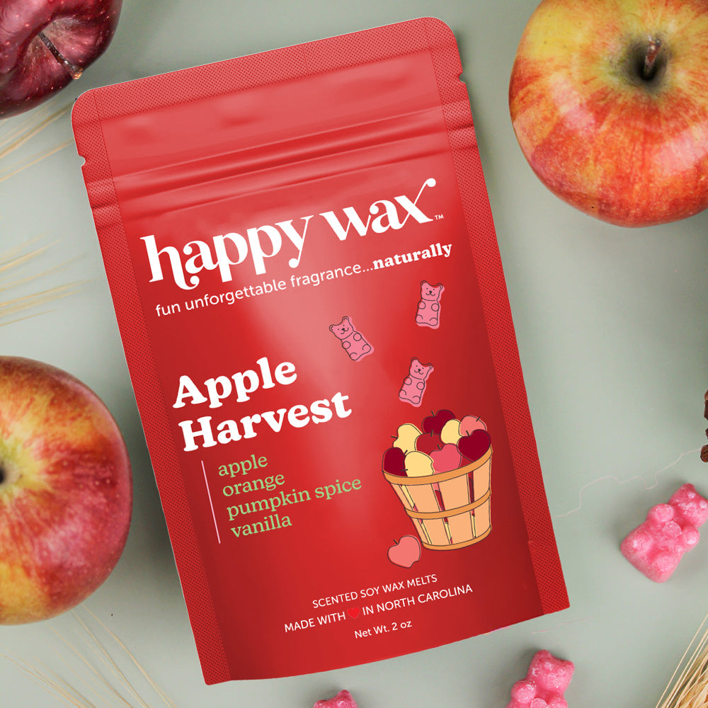 Red pouch of Apple Harvest wax melts surrounded by fresh apples and pink wax melt pieces on a light surface.