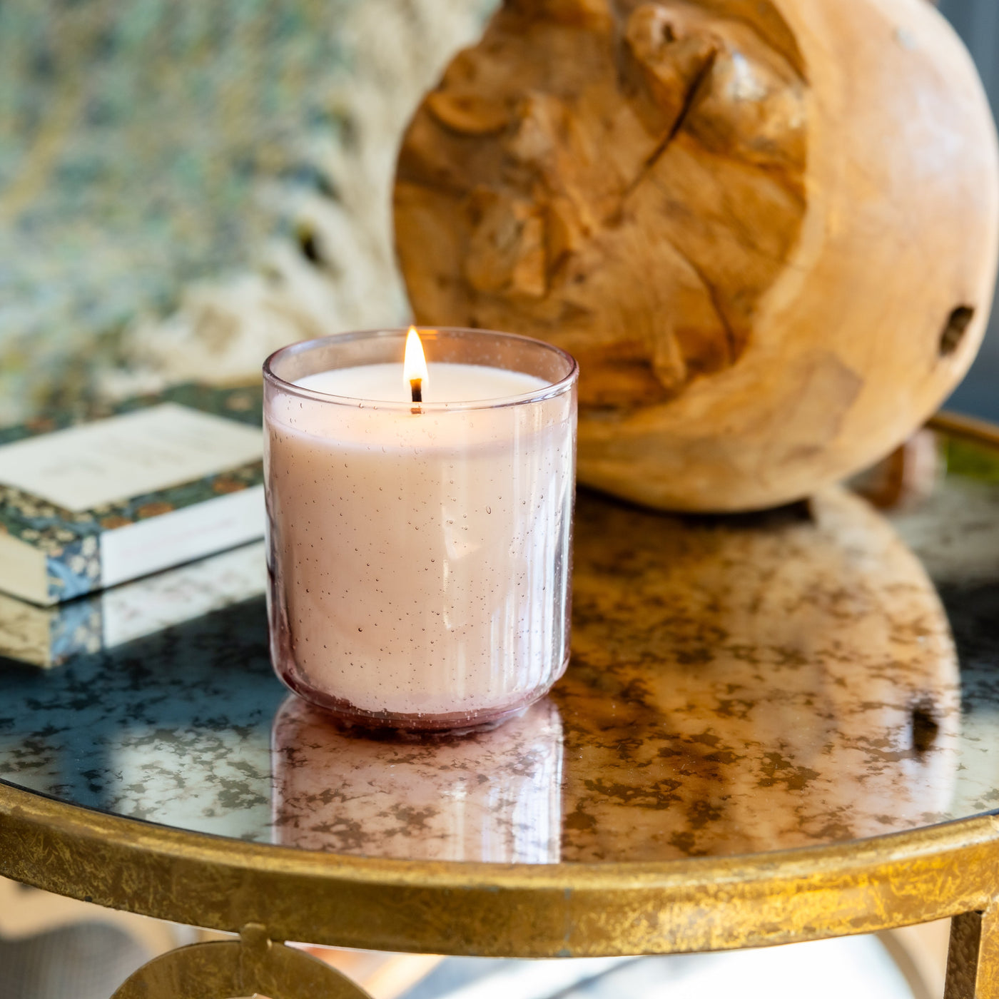 Cream-colored single wick candle in clear glass jar with lit flame, displayed on gold table.