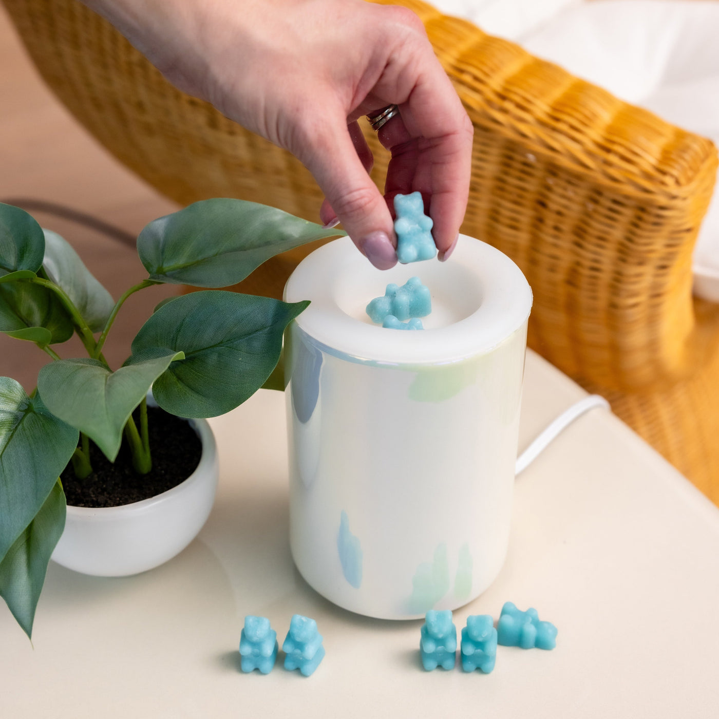 Hand holding blue wax cube above white cylindrical warmer with pastel watercolor design and scattered wax pieces.
