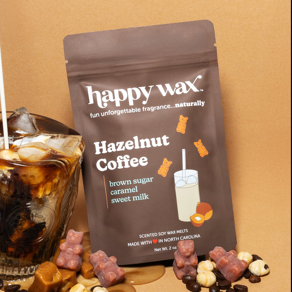 Brown pouch of Happy Wax hazelnut coffee scented soy wax melts displayed with a glass of iced coffee and scattered wax melts.