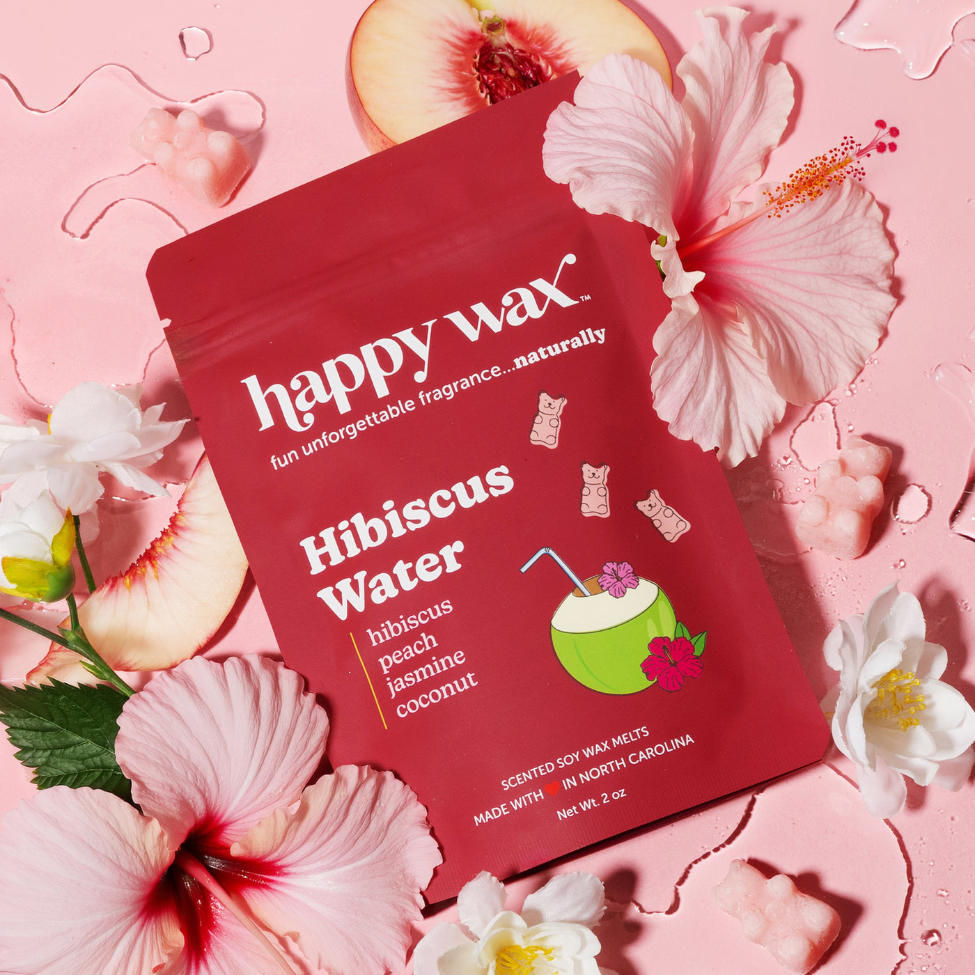 Red pouch of happy wax hibiscus water scented soy wax melts surrounded by pink hibiscus flowers and peach slices on pink back