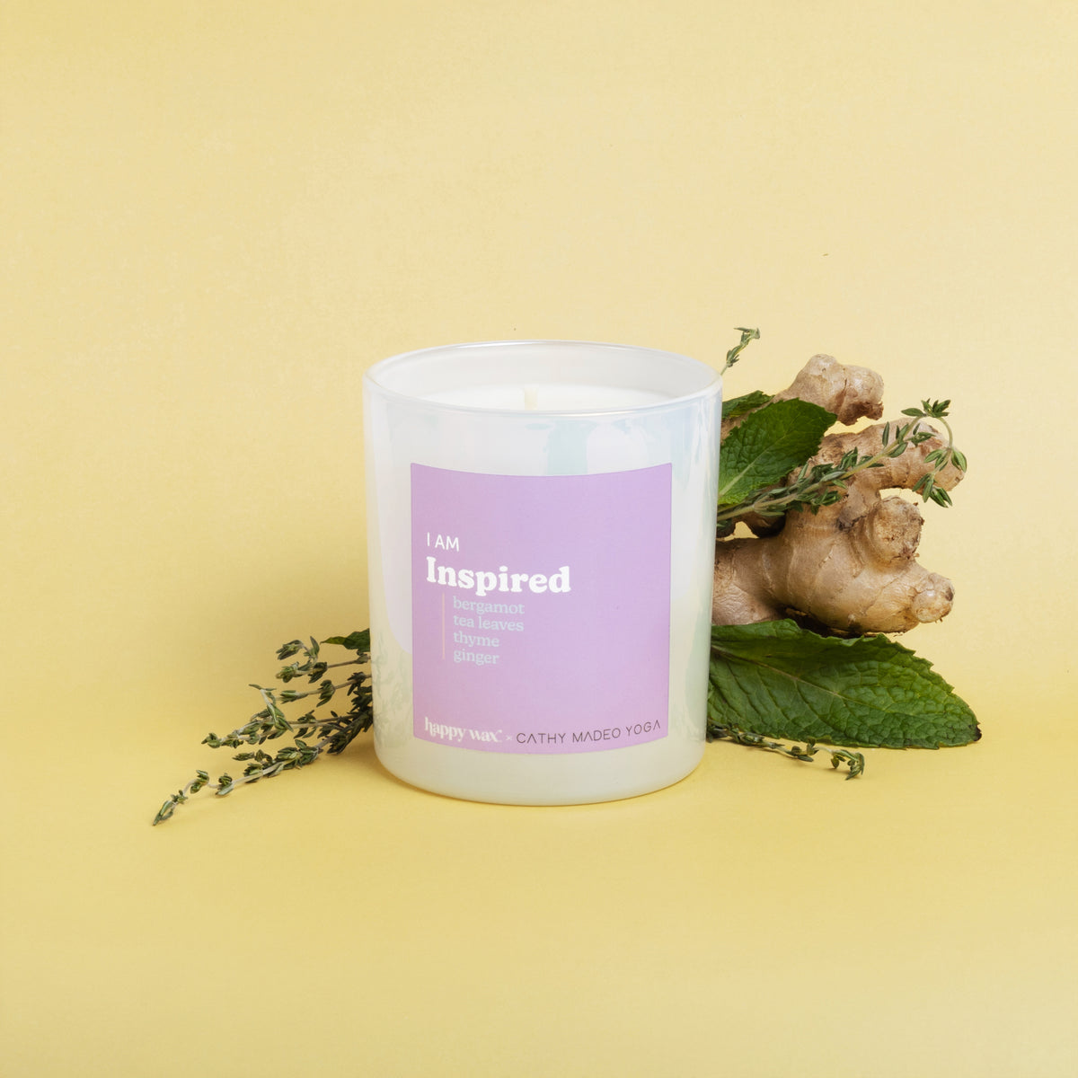 White cylindrical candle with pink label reading "I am Inspired" surrounded by ginger root and green leaves on yellow backgro