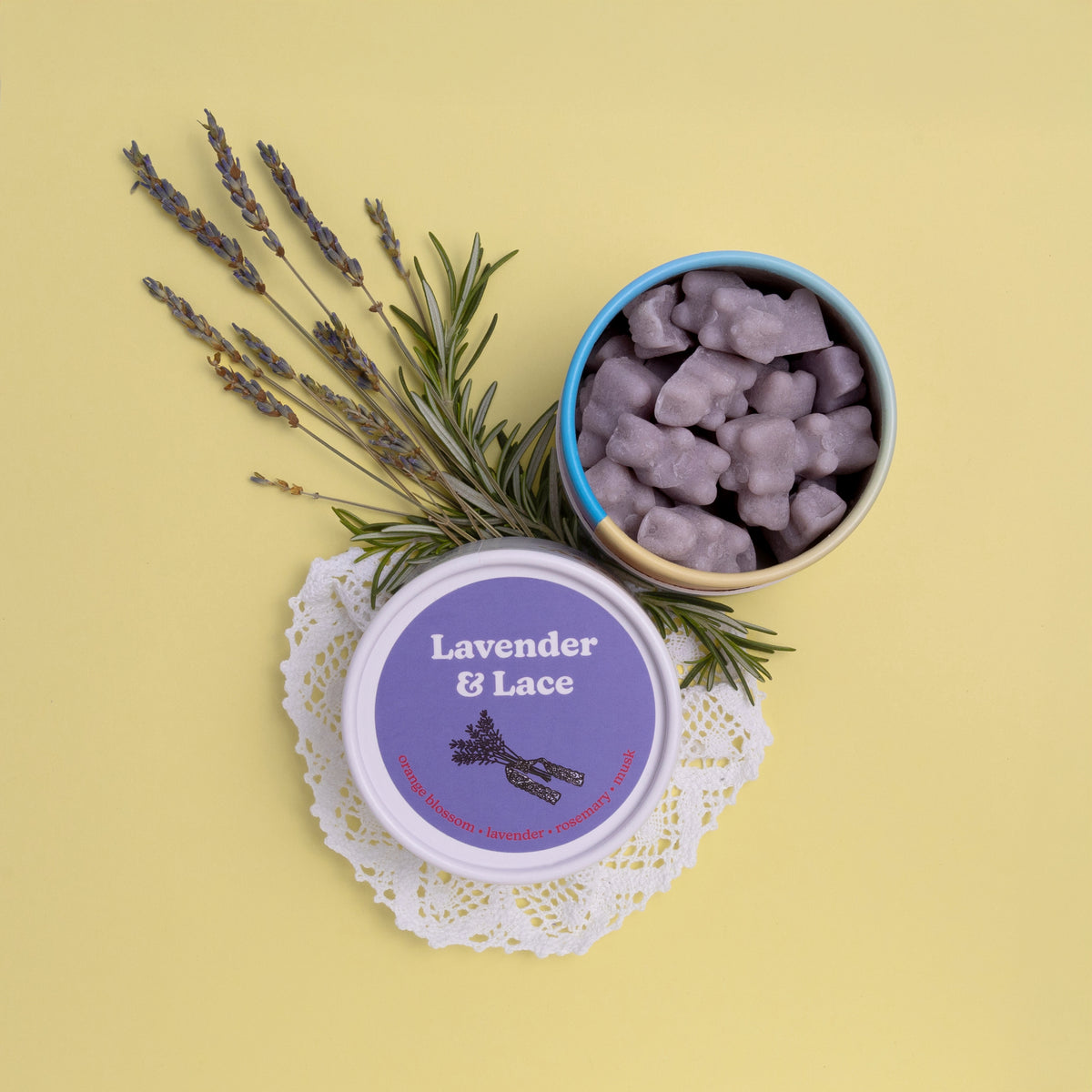 Purple wax melts in blue-rimmed container next to labeled tin, dried lavender sprigs, and white lace doily on yellow backgrou