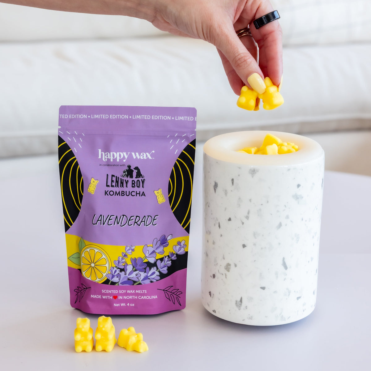 Hand holding yellow wax melt above white speckled wax warmer, with purple "Lavenderade" branded packaging and loose melts dis
