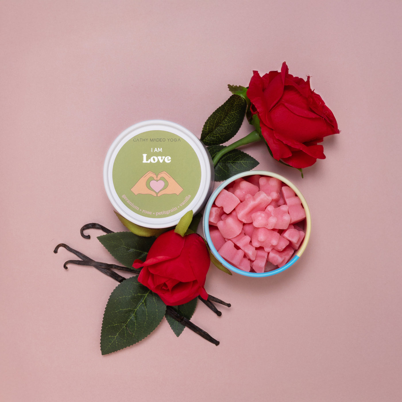 Two wax melt containers with "I am Love" labels surrounded by red roses and vanilla beans on a pink background.