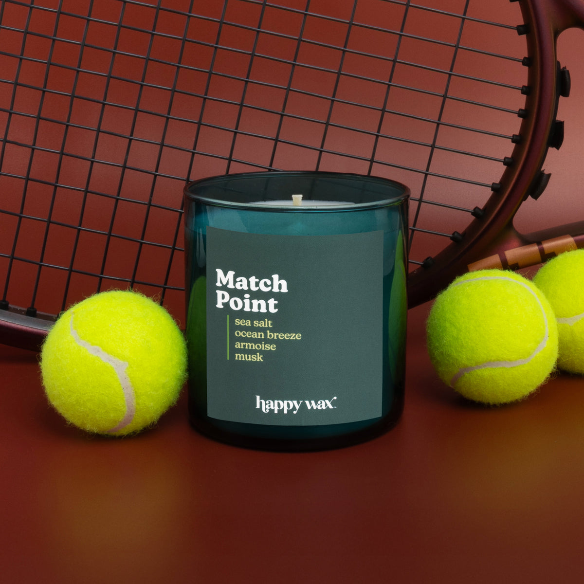 Dark teal glass candle with black lid and single wick, surrounded by tennis balls and racket.