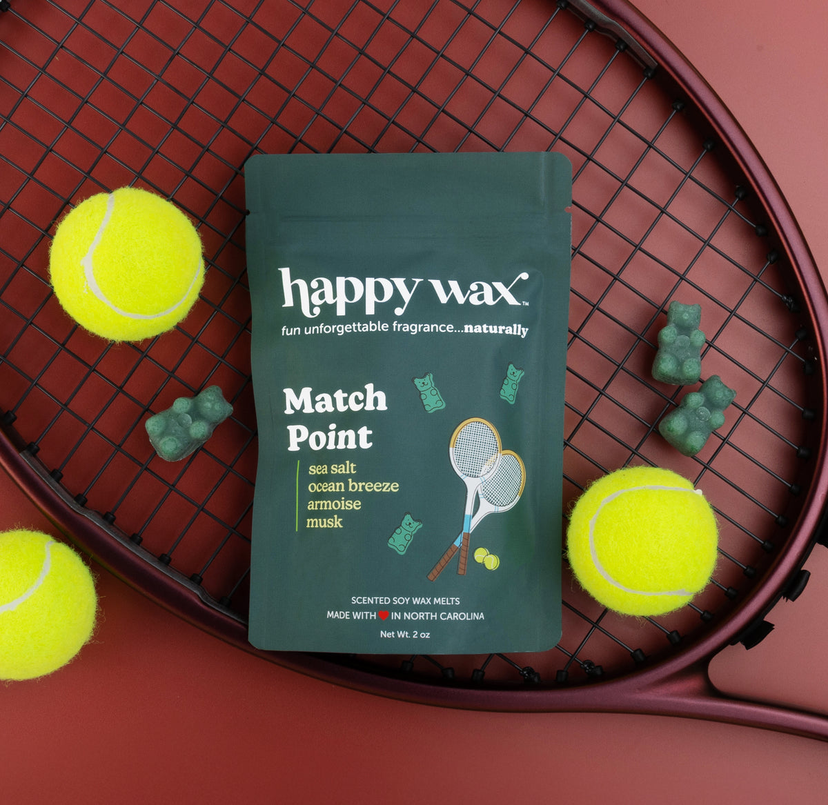 Dark green "Match Point" wax melt package surrounded by yellow tennis balls and green wax melts on a tennis racket.