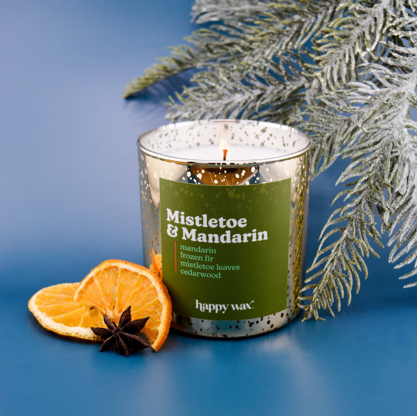 Glass candle with green label, single orange flame, orange slice and star anise beside it, frost-dusted evergreen branches in