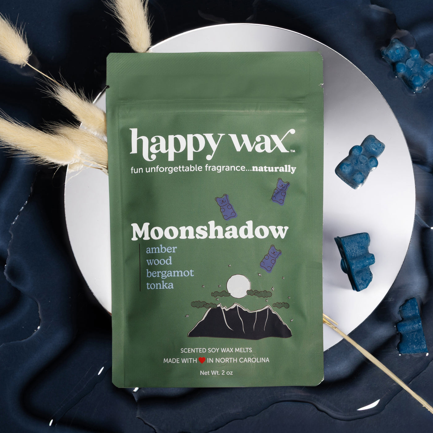 Green Moonshadow wax melt packet on white plate with blue wax melts and pampas grass.