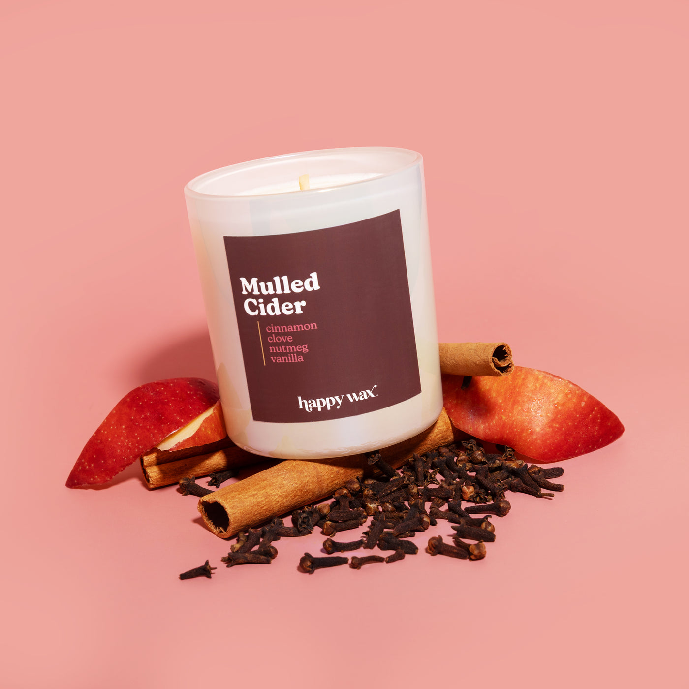White cylindrical candle with dark burgundy label reading "Mulled Cider" surrounded by cinnamon sticks, cloves, and apple sli