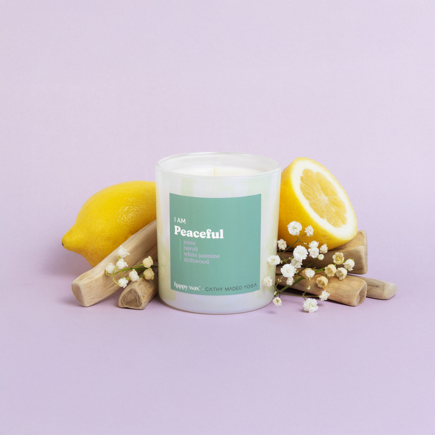 White cylindrical candle with green label surrounded by lemon slices, wooden pieces, and white flowers on purple background.