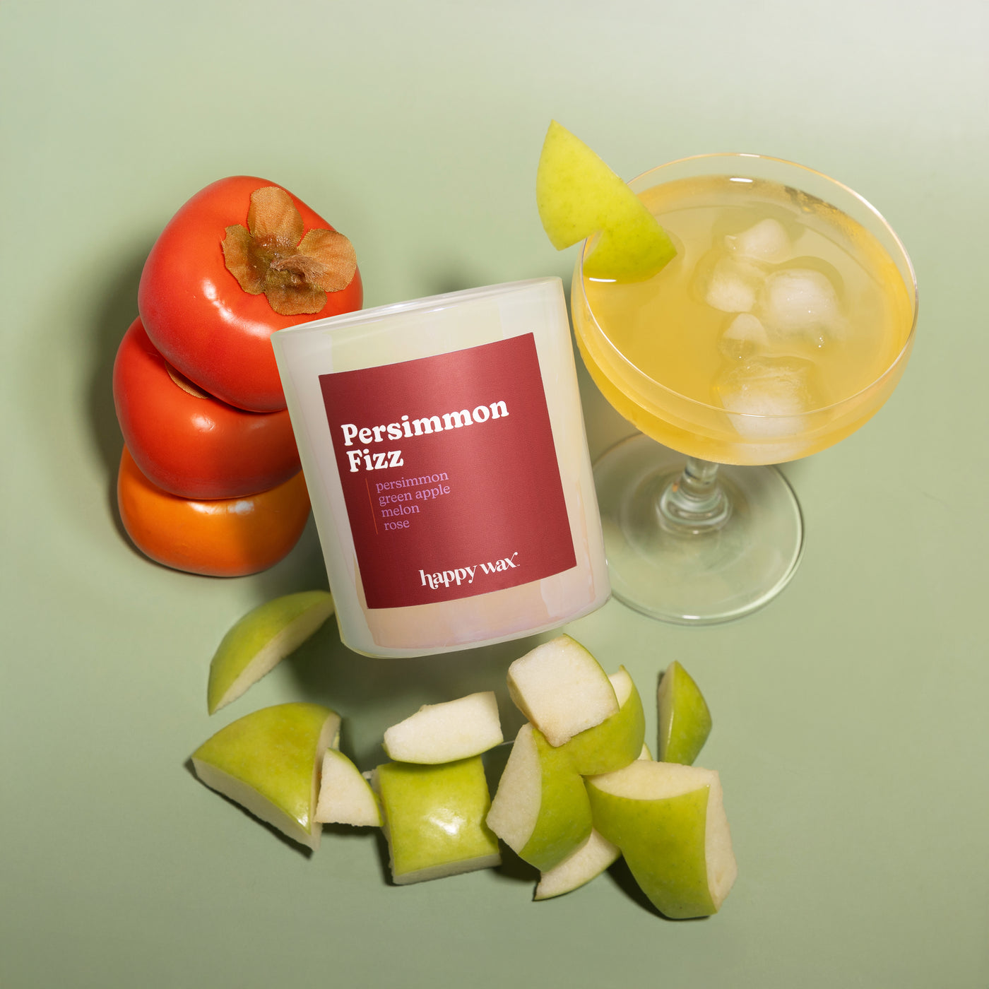 White candle with burgundy label surrounded by persimmons, green apples, and a yellow cocktail with ice.