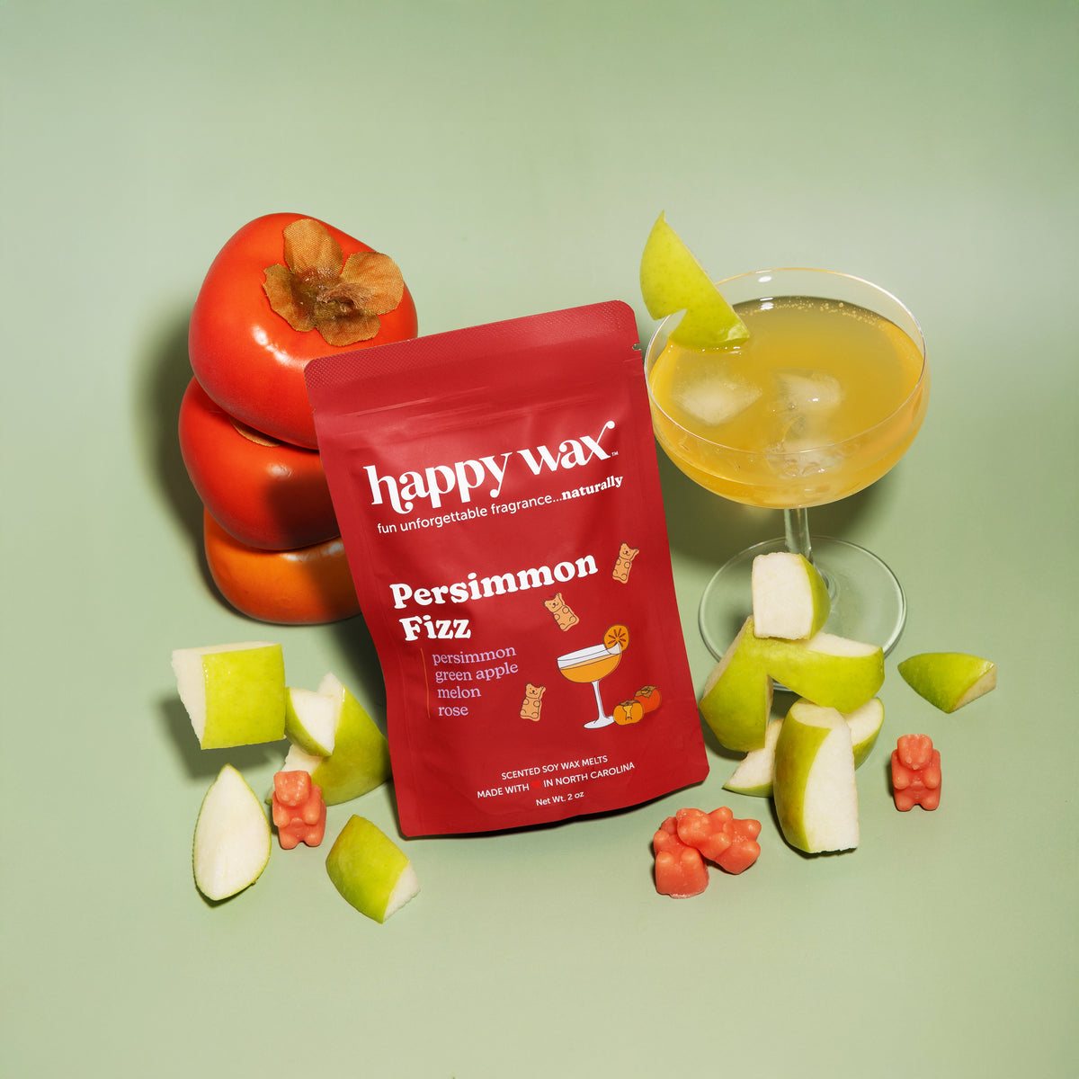 Red "Persimmon Fizz" wax melt packet surrounded by fresh persimmons, green apple slices, a yellow cocktail with ice, and wax 