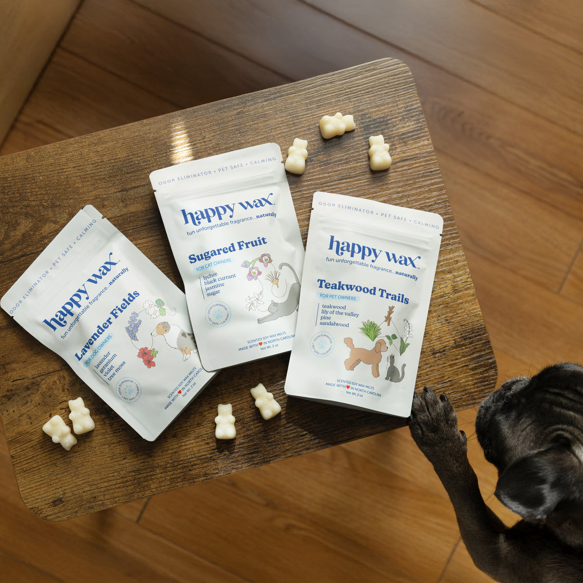 Three Happy Wax pet product pouches in Lavender Fields, Sugared Fruit, and Teakwood Trails scents with wax melts on wooden su