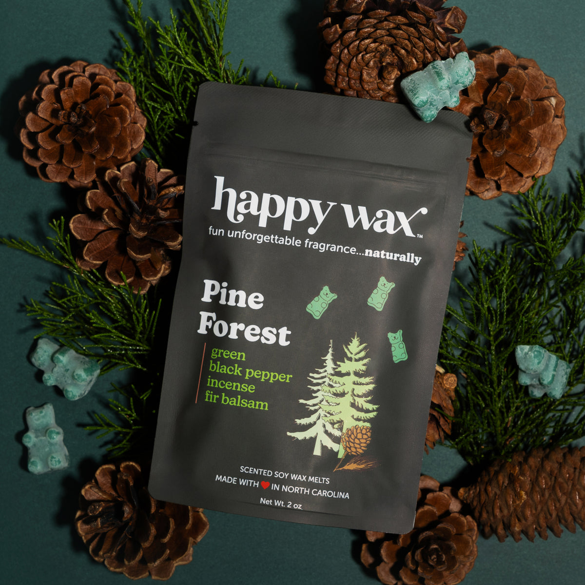 Black pouch of Happy Wax Pine Forest wax melts surrounded by pinecones, evergreen sprigs, and turquoise decorative stones.