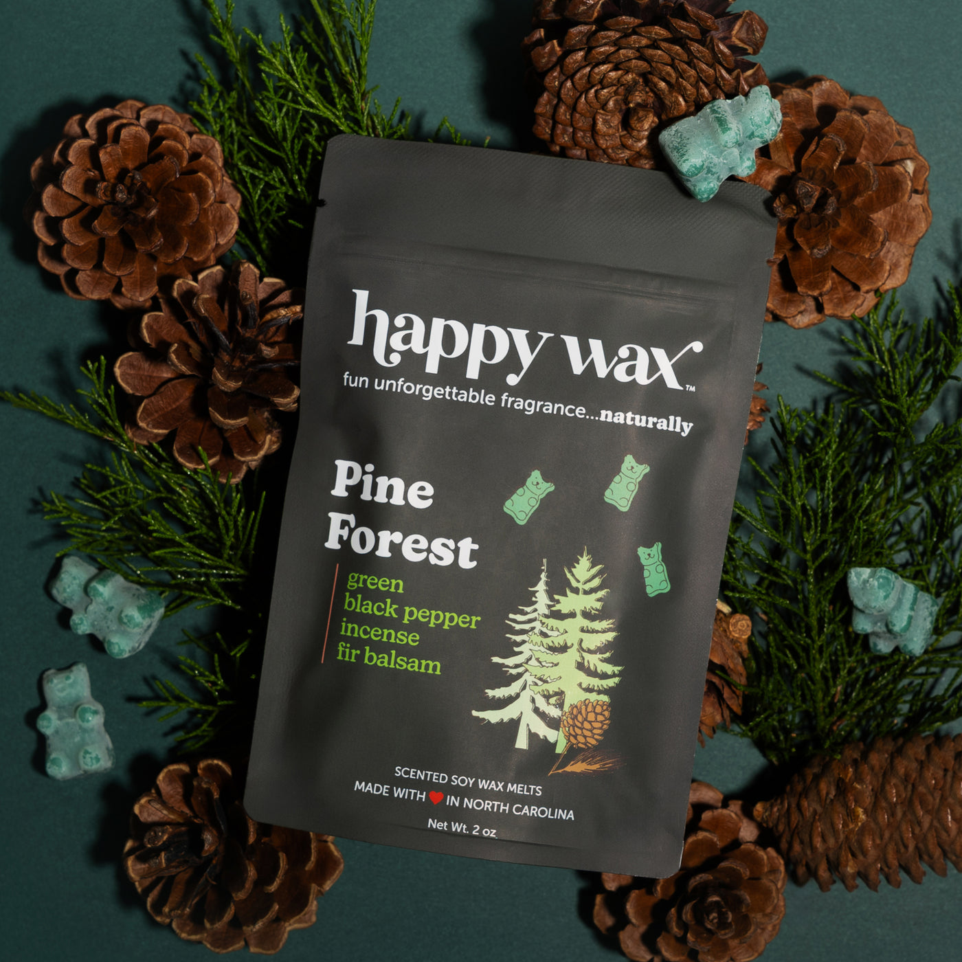Black pouch of Happy Wax Pine Forest wax melts surrounded by pinecones, evergreen sprigs, and turquoise decorative stones.