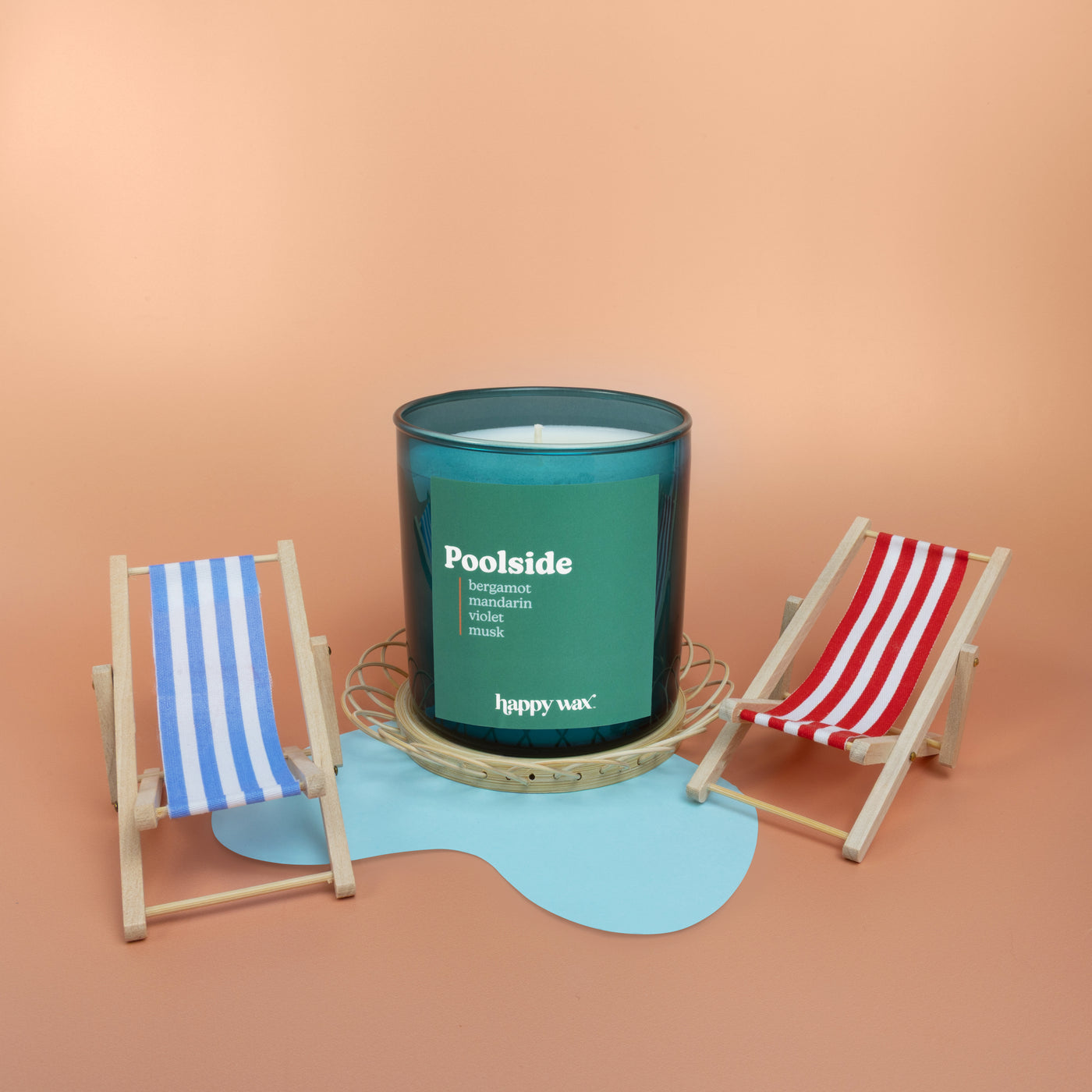 Green glass candle with white single wick surrounded by miniature striped beach chairs and blue wave-shaped base on tan backg
